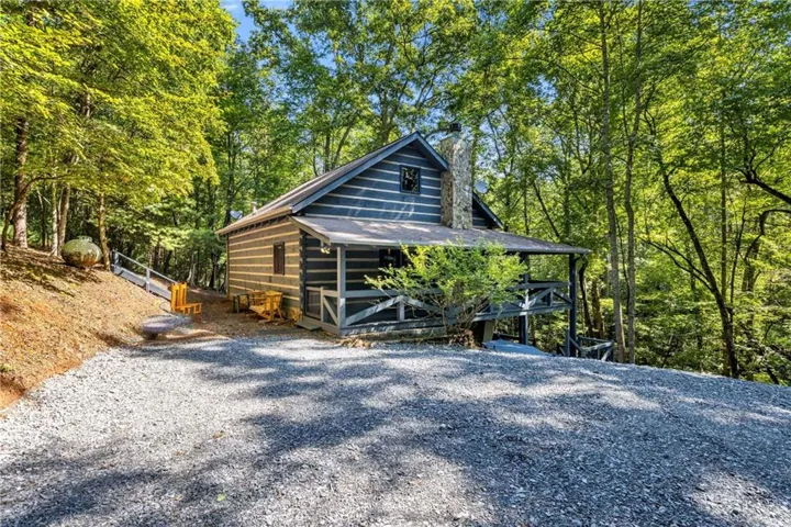 Log cabin exterior with a covered porch, stone chimney, and gravel driveway