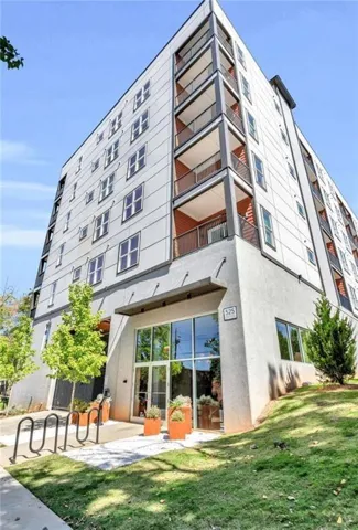 Multi-story building featuring a contemporary facade, individual balconies with metal railings, a ground-level entryway with large glass panels, a prominent address number, and a bicycle rack