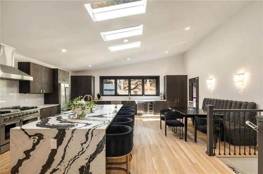 Vaulted ceilings with multiple skylights, custom dark cabinetry, large kitchen island with patterned stone surface, stainless steel appliances, and wood-finish flooring