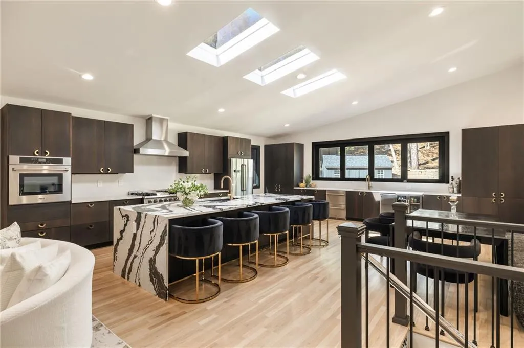 Kitchen featuring dark wood-finish cabinetry, stainless steel appliances, a large island with waterfall-edge countertop, light wood-finish flooring, and multiple skylights