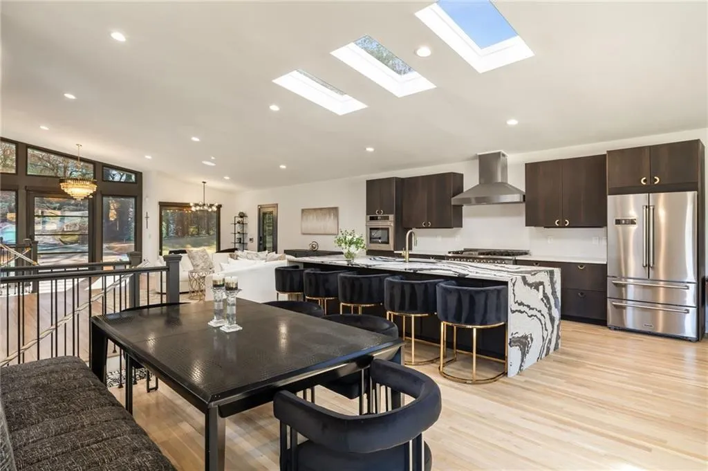 Open-concept living space with light wood-finish flooring, a contemporary kitchen island featuring a waterfall counter, dark wood cabinetry, and multiple skylights
