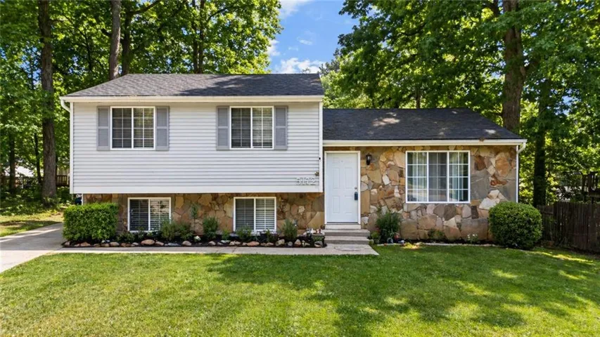 Split-level home with natural stone facade, white vinyl siding, and mature hardwood canopy. Concrete driveway and landscaped entry beds with potted plants along the foundation.