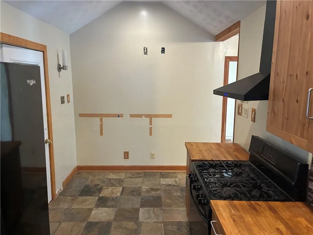 Kitchen featuring butcher block counters, black range with gas cooktop, wall chimney exhaust hood, dark stone finish flooring, and lofted ceiling