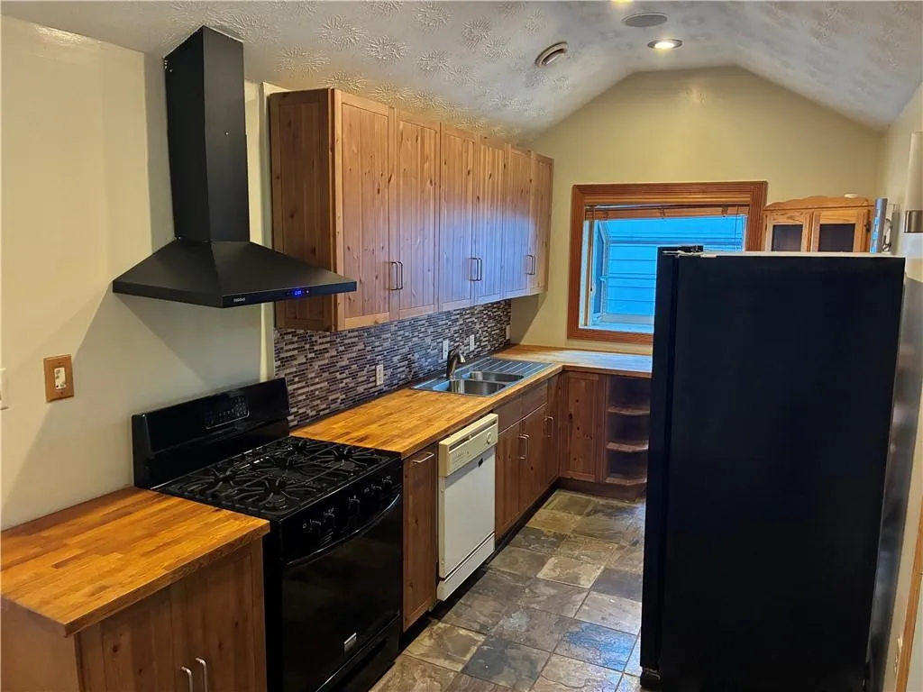 Kitchen featuring black appliances, wall chimney exhaust hood, butcher block counters, brown cabinets, and decorative backsplash