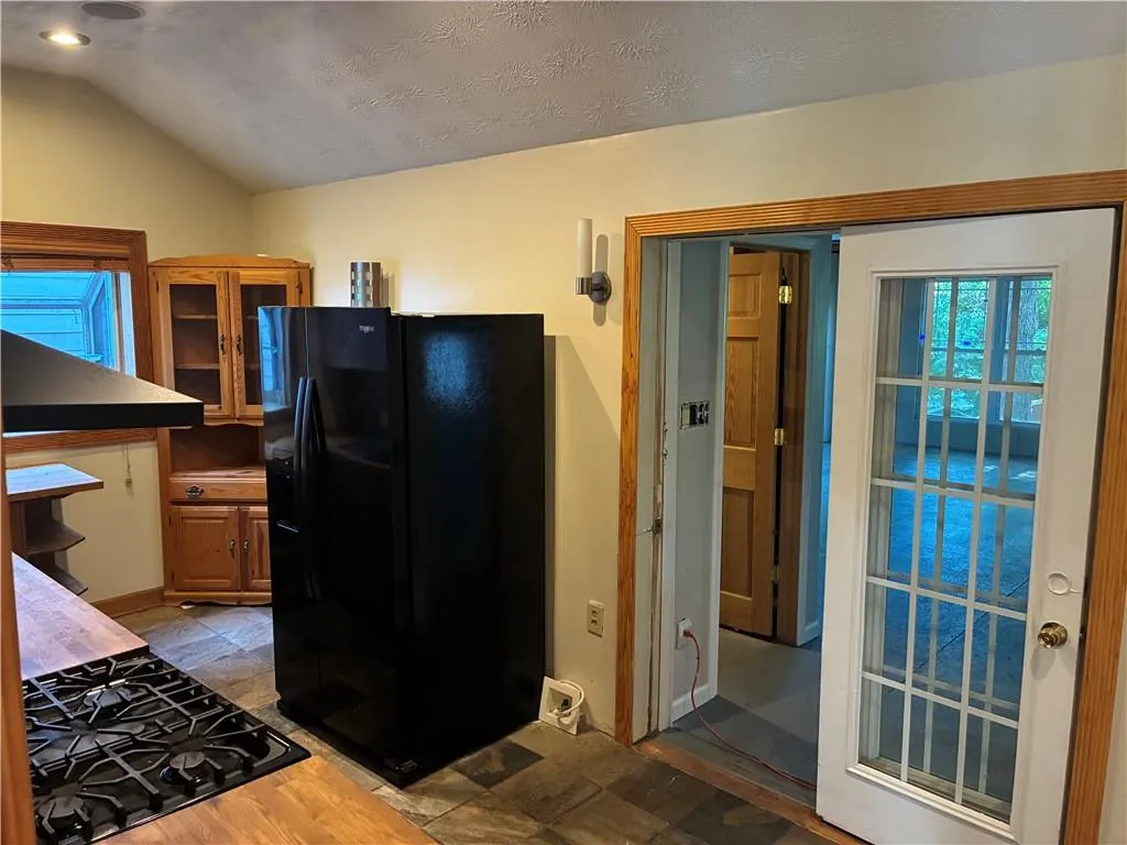 Kitchen with black fridge with ice dispenser, under cabinet range hood, brown cabinetry, glass insert cabinets, and vaulted ceiling