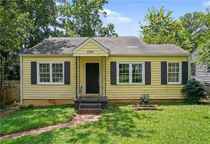 Yellow siding exterior featuring dark shutters, a covered entry with wrought iron railings, and a front yard with mature trees