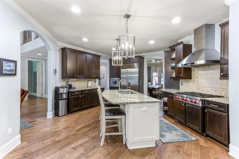 Kitchen with wall chimney exhaust hood, sink, light stone counters, built in appliances, and an island with sink