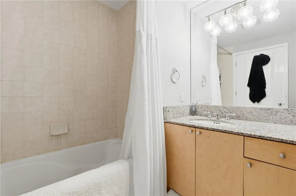 Full-width vanity mirror, light wood-finish cabinetry, granite countertop, undermount sink with a chrome faucet, and a tiled tub surround