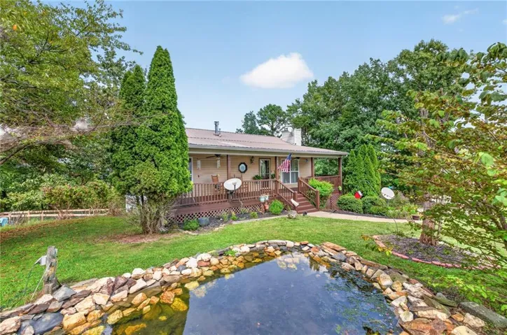 Expansive front yard featuring a rock-lined pond, mature trees, and a walkway leading to a covered porch with wood railings and steps