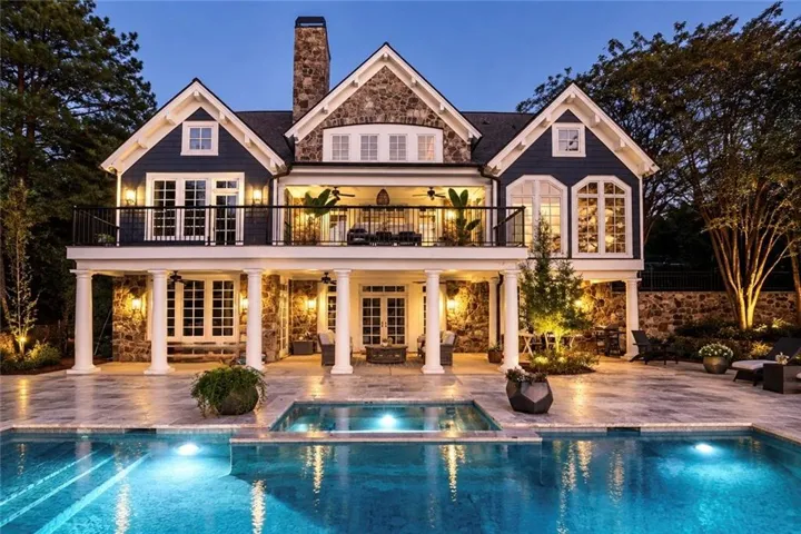 Stone and siding exterior with multiple gables, an upper-level balcony, a covered lower-level patio with columns, and a built-in pool with an integrated spa