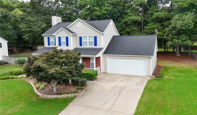 Two-story Colonial featuring a white exterior with blue shutters and brick accents, a two-car garage with a concrete driveway, and a landscaped front yard with mature trees and mulched planting beds set on a spacious lawn