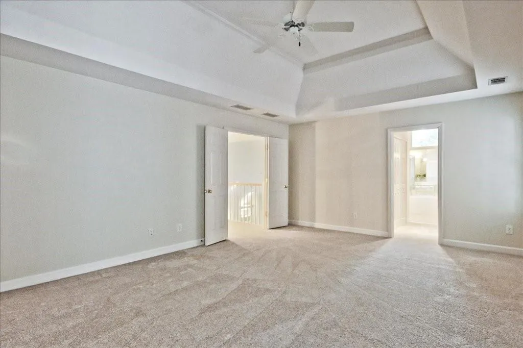 Unfurnished bedroom with a tray ceiling, light colored carpet, ensuite bathroom, and a ceiling fan