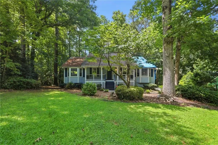 Light blue siding exterior featuring a covered front porch with white railings, dark shutters, a bright blue roof section, and an expansive lawn surrounded by mature trees