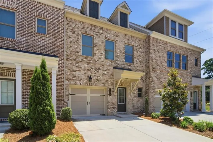 Brick facade townhome featuring a dormer window, covered entry, attached garage, concrete driveway, and mature landscaping