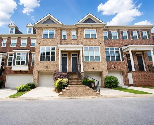 Brick townhouse exterior featuring a two-car garage, prominent front steps with metal railings, a gabled roofline with shingle accent, multiple windows, and manicured shrubbery