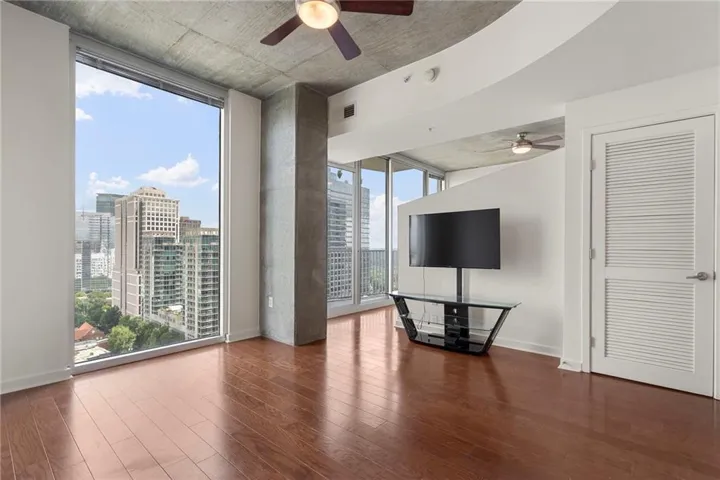 Expansive floor-to-ceiling windows, wood-finish flooring, exposed concrete ceiling, integrated ceiling fans, and a louvered door