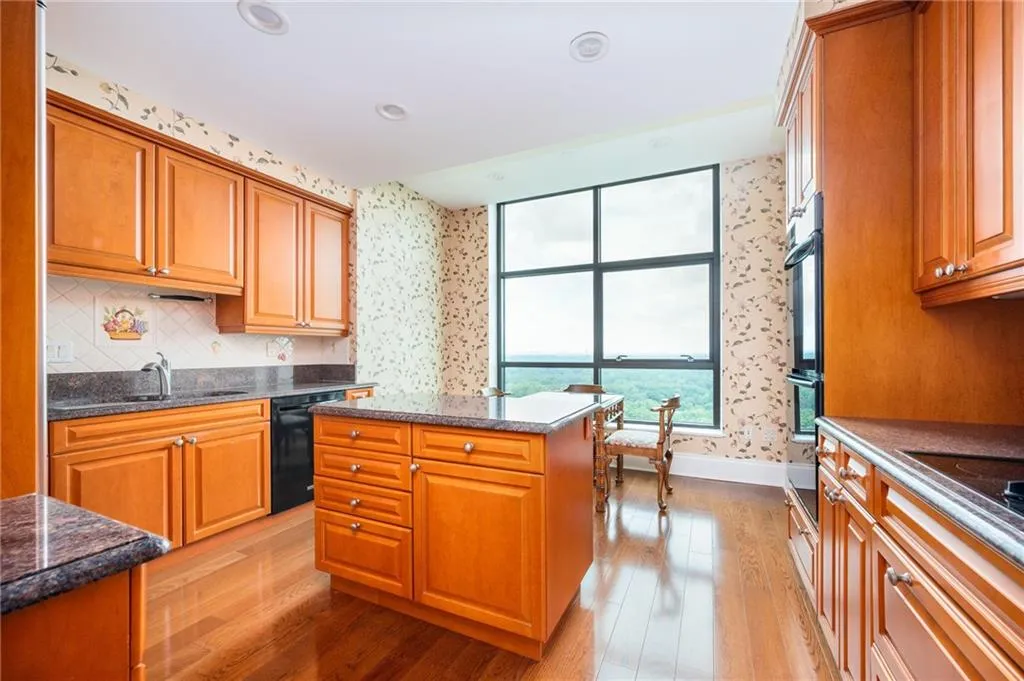Spacious kitchen featuring rich wood cabinetry throughout, stainless steel appliances, granite countertops, a centered island with seating, wood-finish flooring, and abundant natural light from large multi-pane windows overlooking the surrounding landscape