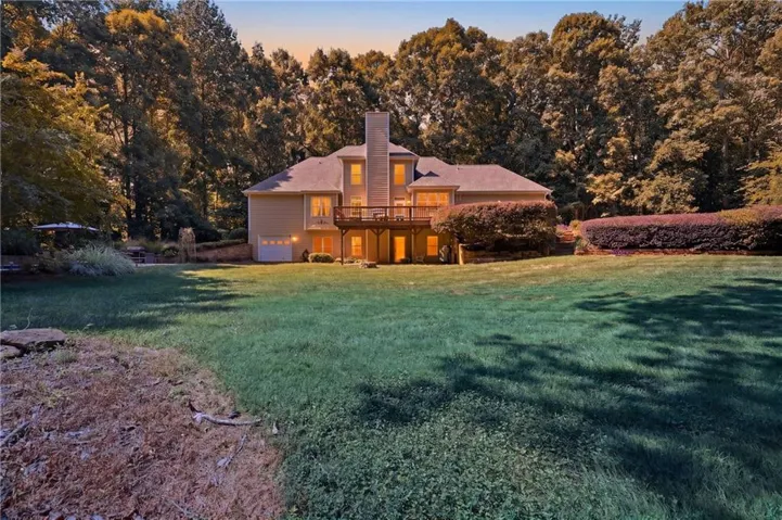 Expansive green lawn with a two-story residence featuring a wood-finish deck, prominent chimney, and mature perimeter landscaping