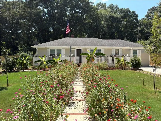 Charming single-story residence with a welcoming front porch and white railings, set back from a landscaped approach featuring flowering shrubs and mature trees
