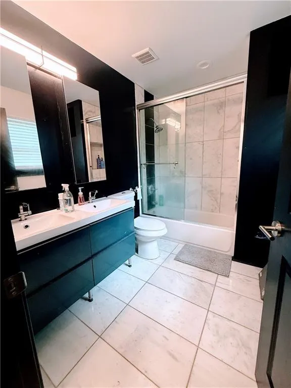 Full bath with double vanity, bath / shower combo with glass door, toilet, and tile patterned floors