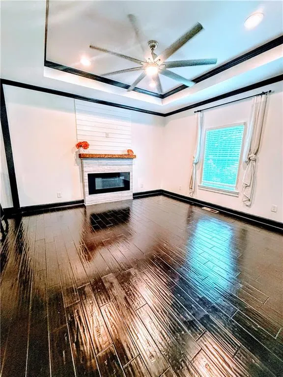 Unfurnished living room with a ceiling fan, a tray ceiling, wood-type flooring, a glass covered fireplace, and baseboards