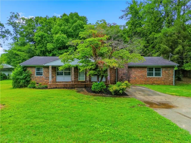 Brick exterior residence featuring a covered front porch with white columns, a dark shingle roof, a manicured front lawn, and mature trees providing natural screening