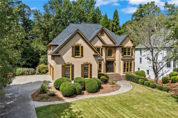 Stucco exterior with stone accents, dark-shingle roof, wood shutters, concrete driveway, and established landscaping
