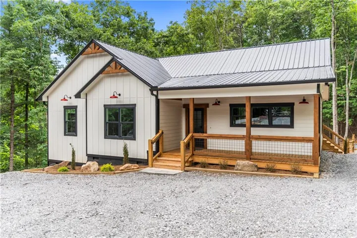 Modern farmhouse exterior featuring vertical white siding, black-framed windows, a covered porch with natural wood railing, a metal roof, and exposed wood rafter tails