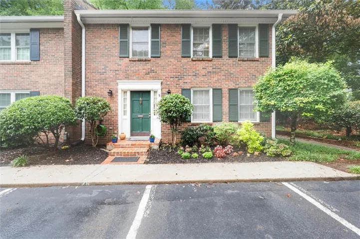 Brick facade townhome featuring a vibrant green entry door with white trim, dark green shutters, and a well-maintained front landscaping with mature plantings