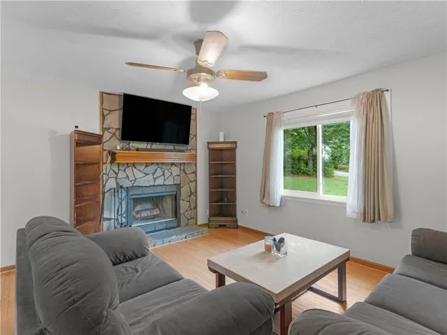 Stone-clad fireplace with wood mantel, wood-finish flooring, ceiling fan with light fixture, and window with outdoor greenery