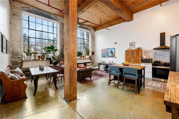 Exposed brick walls, expansive industrial-style windows, raw wood ceiling beams, polished concrete flooring, and a contemporary kitchen with subway tile backsplash