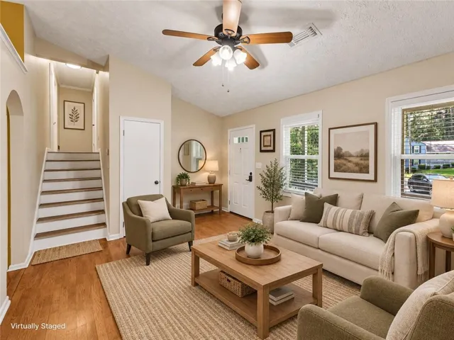 Wood-finish flooring, vaulted ceiling, ceiling fan with integrated lighting, multiple windows with blinds, and a white paneled entry door