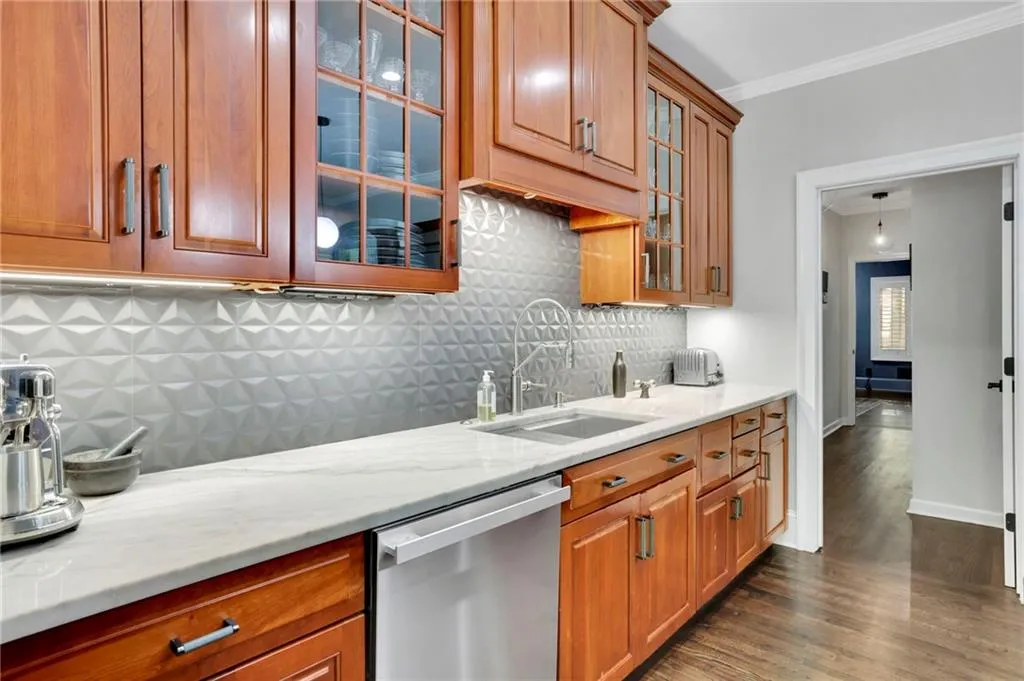 Kitchen with dark wood-style floors, stainless steel dishwasher, brown cabinetry, backsplash, and light stone counters
