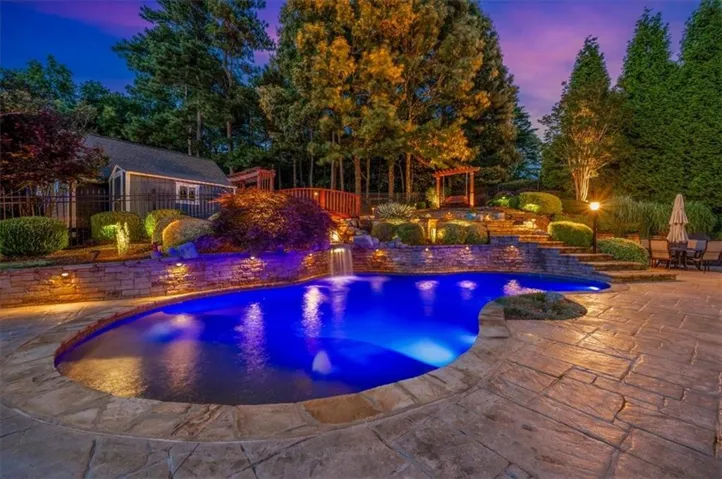 Freeform swimming pool with a waterfall feature, stamped concrete patio, tiered landscaping with integrated lighting, and a wooden gazebo