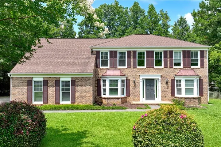 Brick-front Colonial-style residence featuring a two-story design, dual bay windows with copper awnings, a dark paneled front door with white surround, and a manicured lawn with mature landscaping