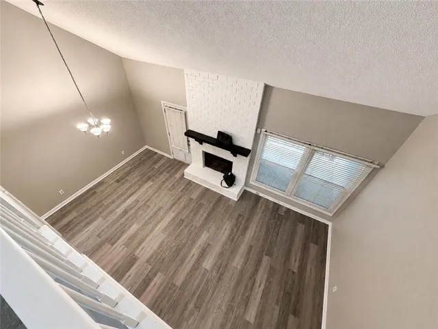Vaulted ceilings with a suspended light fixture, wood-finish flooring, a white brick fireplace with a dark mantle, and two windows with blinds