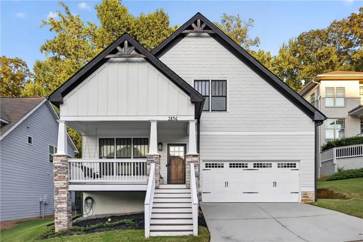 Modern farmhouse exterior featuring a white fiber cement board facade with black architectural accents, a covered front porch with white railings and stone pillar supports, a two-car garage with paneled white doors, and mature landscaping with established trees framing the property