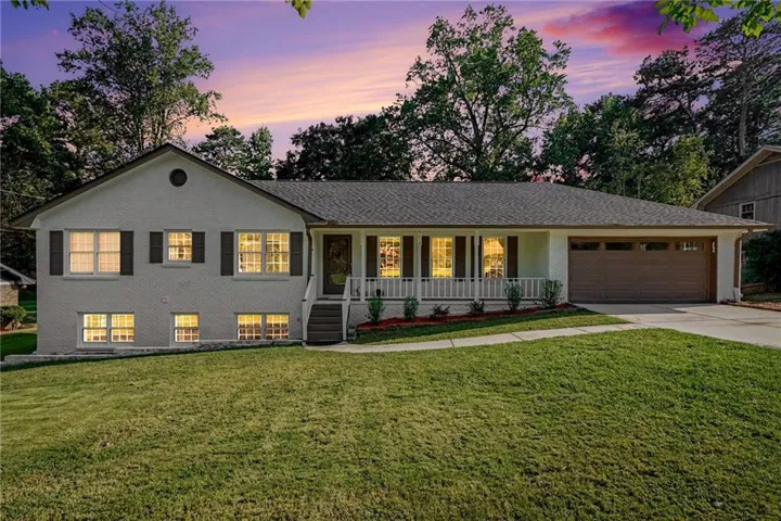 White exterior with dark shutters, covered front porch, attached garage with a wood-finish door, and a shingle roof