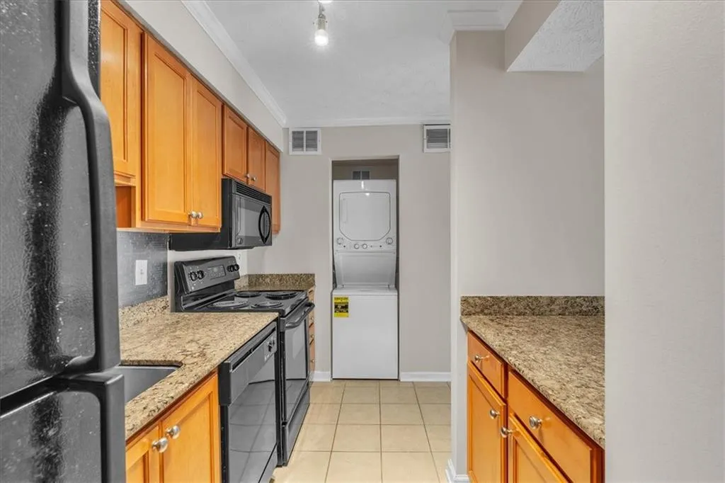 Kitchen with black appliances, light stone countertops, crown molding, stacked washer / dryer, and light tile patterned floors