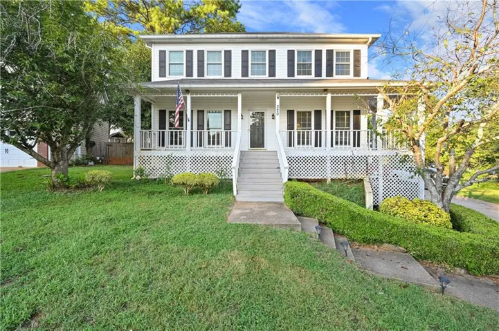 Two-story exterior with white siding, black window shutters, a full-width front porch with white railing, and a sloped front yard with mature trees