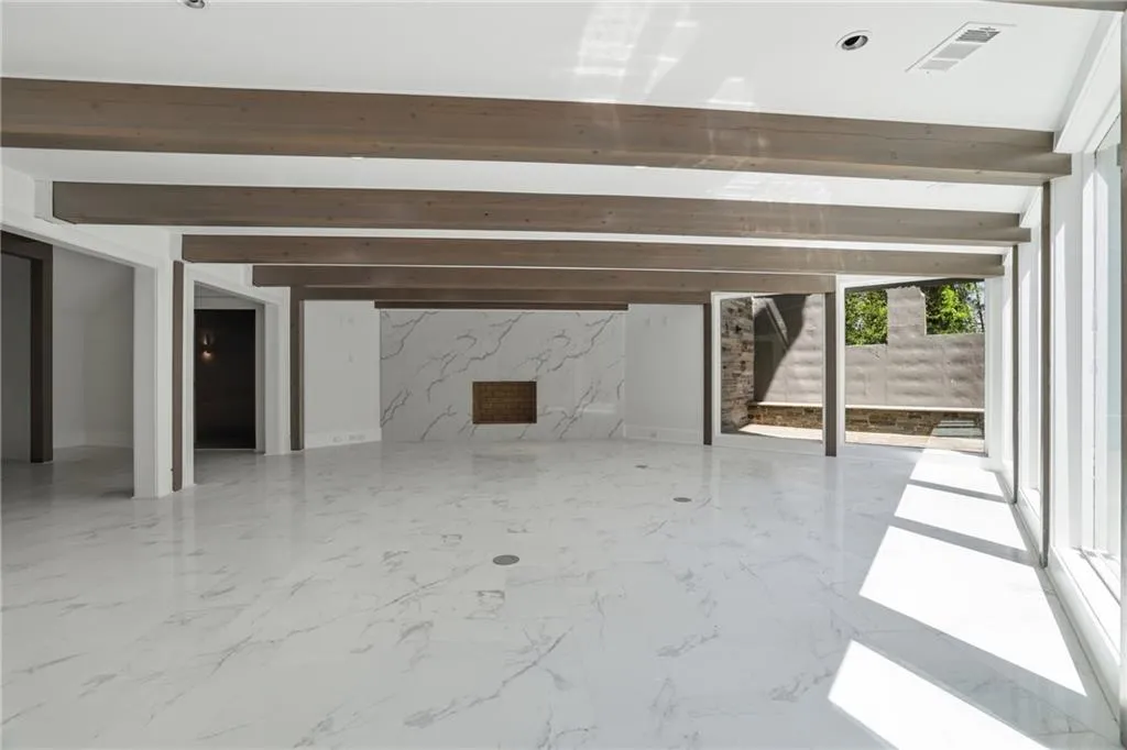 Spacious interior featuring a white ceiling with exposed wood-finish beams, large format white marble-look tile flooring, and a fireplace with a white marble-look surround and brick firebox