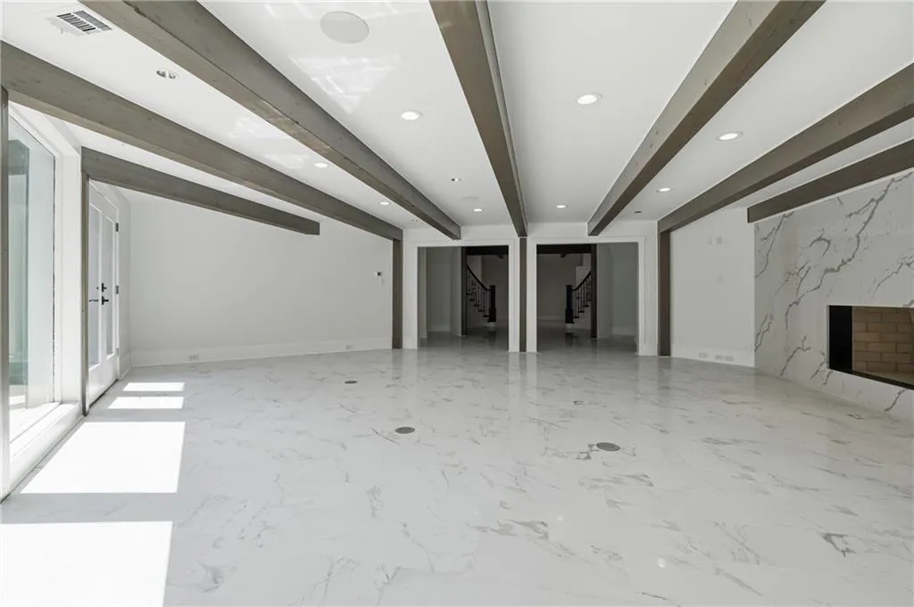 Expansive interior space featuring a large format white tile floor, a fireplace with a stone-look surround, exposed wood beams, recessed lighting, and a glass paneled door
