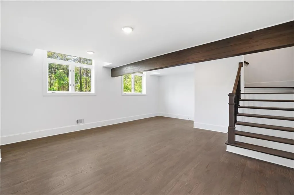 Spacious room featuring wood-finish flooring, two large windows, recessed lighting, a dark wood beam, and a staircase with dark wood treads and a complementary handrail