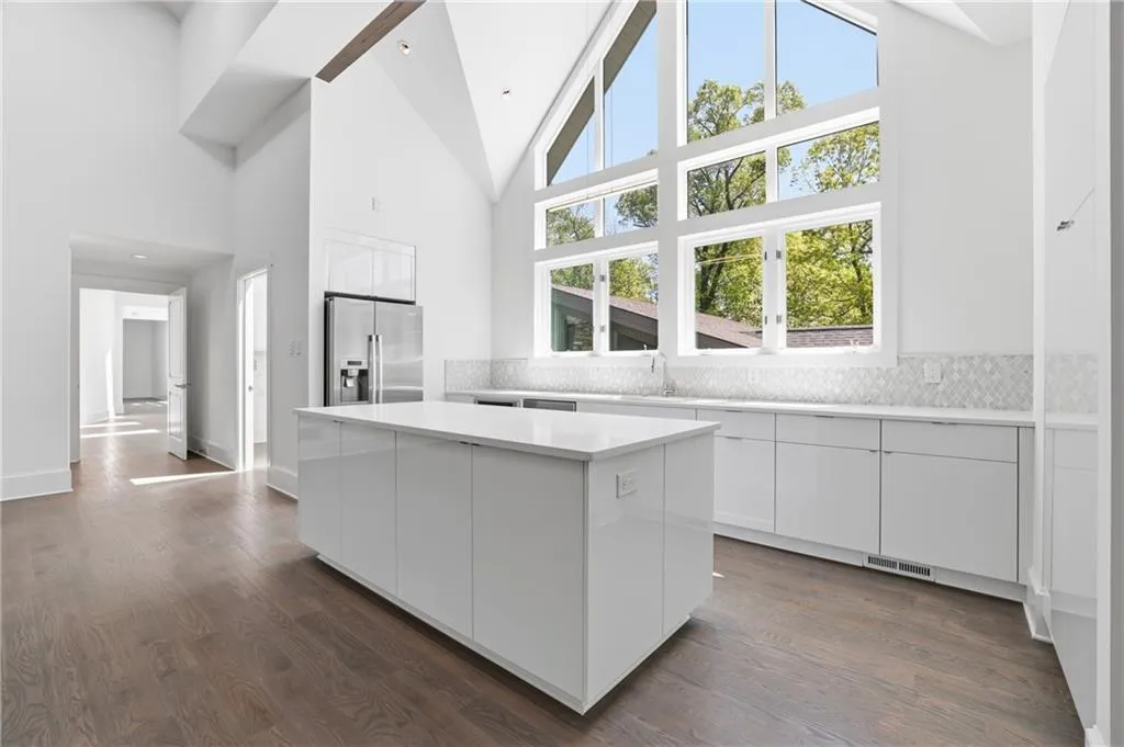 Contemporary kitchen featuring vaulted ceilings, a large central island with a solid surface countertop, white cabinetry, stainless steel refrigerator, and a window wall overlooking mature trees