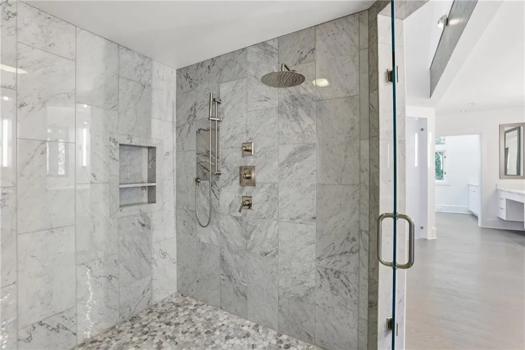 Spacious walk-in shower featuring full-height stone tile walls, a rain shower head, a handheld shower fixture, a built-in shower niche, and a pebble-finish floor
