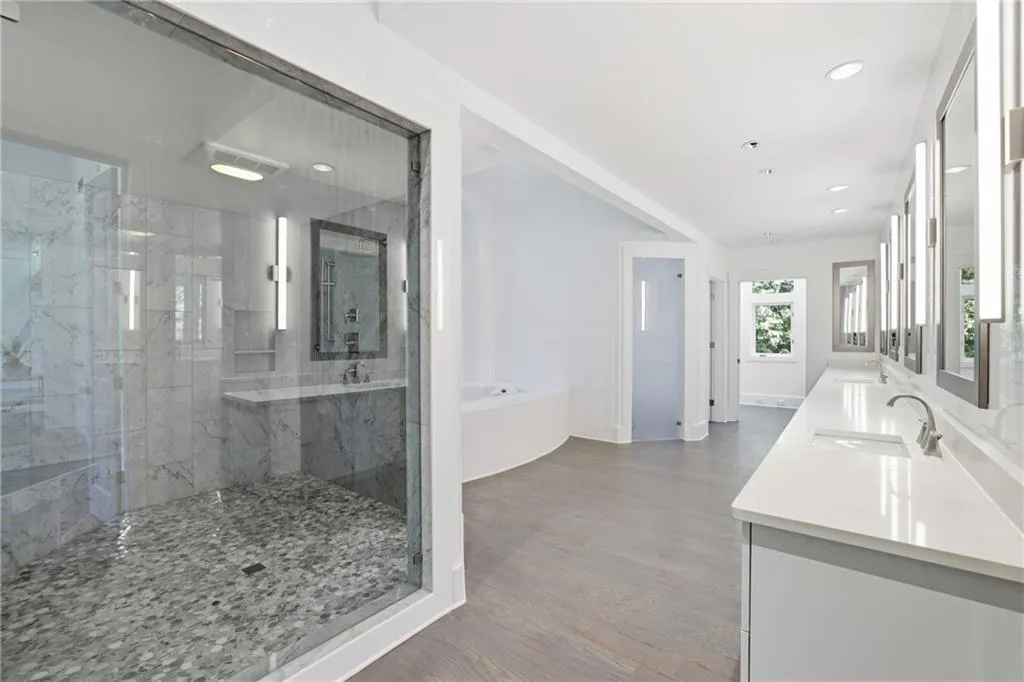Spacious bathroom featuring a glass-enclosed shower with pebble flooring and marble-finish wall tiling, a built-in soaking tub, a multi-sink vanity with light countertops, and wood-finish flooring