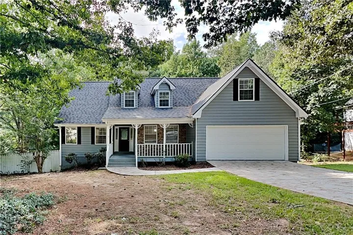 Gray siding exterior with white trim, covered front porch with stone accents, two dormer windows, attached garage, and paved driveway