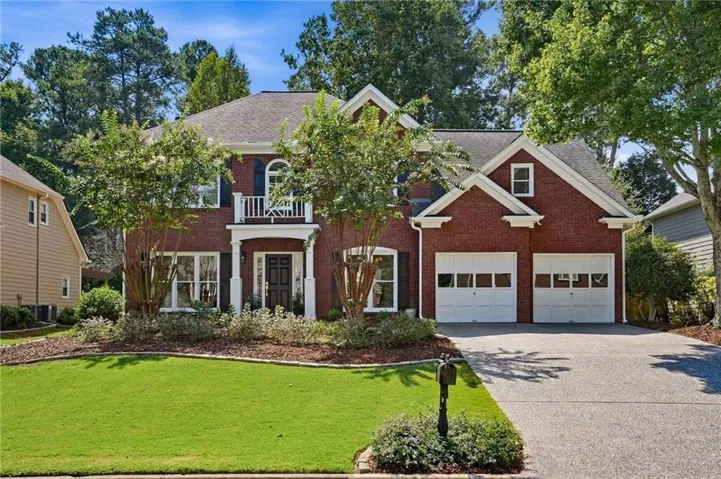 Red brick exterior with white trim, a covered front entry with white columns, a second-story balcony with white railing, and a two-car garage with panel doors