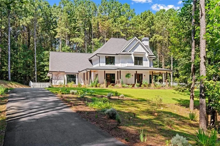 Modern farmhouse architecture with white vertical siding, a dark pitched roof, a covered front porch with wood columns, a stone chimney, and a paved driveway