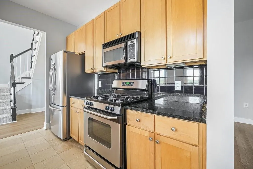 Kitchen featuring light tile flooring, stainless steel appliances, dark stone counters, light brown cabinetry, and tasteful backsplash Kitchen featuring light tile flooring, stainless steel appliances, dark stone counters, light brown cabinetry, and tasteful backsplash